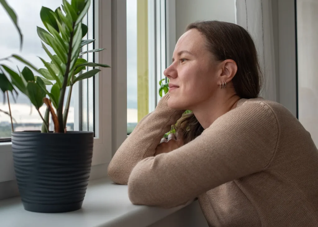Woman handling a breakup by acting compassionately and thinking while sitting by her window with green plants