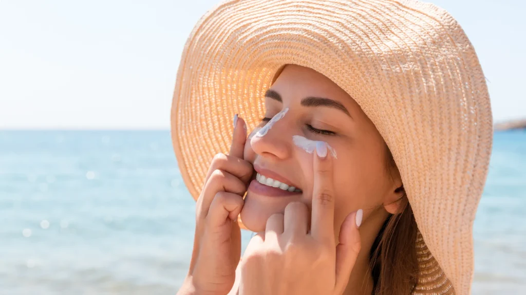 Why is sunblock important? Woman wearing a wide brim sun hat applying sunscreen to her face