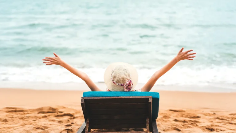 Why Vacations Are Important - Woman sitting on a beach chair in the sand close to the ocean water with her arms in the arm
